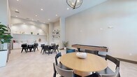 Located in North Port, Florida, this single-floor amenity space is thoughtfully designed to serve both social and wellness needs. The layout is divided into two distinct areas that together create a cohesive, modern environment.

The dining and social area exudes a contemporary aesthetic, featuring soaring ceilings that lend an open, airy feel to the room. Light-toned large-format tile flooring runs throughout, complementing the neutral color palette of soft whites and warm grays on the walls. Multiple seating arrangements — including a round wood-top table with mixed chairs and a marble-top rectangular table with black mid-century style chairs — offer flexible gathering options. A built-in kitchenette with white cabinetry, open shelving displaying curated décor, and stainless steel appliances anchors one end of the room. A wood shuffleboard table adds a recreational touch, while an elegant chandelier and recessed lighting illuminate the space warmly. Arched architectural details and tall double doors add character and grandeur.

The exercise room is generously sized and bathed in natural light through a row of large windows, offering views of the surrounding greenery. High ceilings and a ceiling fan keep the space comfortable. Carpeted flooring provides cushioning for workouts, and the room is equipped with a wide array of cardio machines, a multi-station cable machine, free weights, and exercise balls, making it a comprehensive fitness facility.