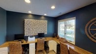 This single-story home office is designed to serve as a fully functional and visually striking workspace. The room is defined by its deep navy blue walls, which create a bold, focused atmosphere well-suited for productivity. A textured popcorn ceiling is outfitted with recessed lighting that provides consistent, even illumination throughout the space.

The centerpiece of the room is a built-in L-shaped desk with a warm butcher block wood surface, supported by white cabinetry below that offers ample storage. The wraparound configuration accommodates multiple workstations, making the space practical for shared or multi-purpose use. Woven rattan chairs complement the natural wood tones of the desk, adding a casual yet refined touch to the overall aesthetic.

A large window fitted with white horizontal blinds anchors one wall, allowing natural light to filter in while offering a view of the surrounding greenery outdoors. Adjacent to the window, additional cabinet storage continues the built-in design theme.

A prominent textural wall art piece—composed of interlocking natural elements in neutral tones—serves as a dramatic focal point on the main wall, adding visual depth against the dark backdrop. Light tile flooring grounds the room and contrasts the richness of the walls, completing a well-balanced and purposeful office environment.