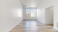 This single-floor unit presents a clean, modern aesthetic defined by crisp white walls, light wood-tone flooring, and an open, airy layout. The main living area is particularly striking, featuring notably tall ceilings and oversized industrial-style grid windows that flood the space with abundant natural light, creating a bright and expansive atmosphere. A built-in shelving unit and a skylight-style ceiling fixture further enhance the living area's functionality and openness.

The bedroom areas continue the light-filled theme, with one room featuring a tall, vertically oriented grid window that draws the eye upward, emphasizing the generous ceiling height. Closets are accessible from both the bedrooms and the hallway, offering ample storage throughout. A long, well-lit corridor connects the rooms, maintaining a clean and organized flow.

The galley-style kitchen is efficiently designed, featuring white cabinetry, dark stone-look countertops, a subtle tile backsplash, stainless steel appliances including a refrigerator and electric range, and marble-style flooring that ties cohesively to the bathroom. The bathroom maintains the same elegant marble-style tile flooring, complemented by a white vanity with gold hardware, a full soaking tub with surrounding shelving, and a bright, all-white finish throughout.
