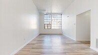 This single-floor unit presents a clean, modern aesthetic defined by crisp white walls, light wood-tone flooring, and an open, airy layout. The main living area is particularly striking, featuring notably tall ceilings and oversized industrial-style grid windows that flood the space with abundant natural light, creating a bright and expansive atmosphere. A built-in shelving unit and a skylight-style ceiling fixture further enhance the living area's functionality and openness.

The bedroom areas continue the light-filled theme, with one room featuring a tall, vertically oriented grid window that draws the eye upward, emphasizing the generous ceiling height. Closets are accessible from both the bedrooms and the hallway, offering ample storage throughout. A long, well-lit corridor connects the rooms, maintaining a clean and organized flow.

The galley-style kitchen is efficiently designed, featuring white cabinetry, dark stone-look countertops, a subtle tile backsplash, stainless steel appliances including a refrigerator and electric range, and marble-style flooring that ties cohesively to the bathroom. The bathroom maintains the same elegant marble-style tile flooring, complemented by a white vanity with gold hardware, a full soaking tub with surrounding shelving, and a bright, all-white finish throughout.