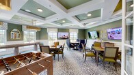 Located at 1500 Laurel Crossing Pkwy NE in Buford, Georgia, this single-floor game and activity room is a well-appointed communal space designed for leisure and social engagement. The room's most striking architectural feature is its coffered ceiling, finished in a muted sage green and crisp white, which lends a refined, upscale character to the space. Gold pendant light fixtures complement the warm tones throughout, while recessed lighting ensures the room is well-illuminated at all times.

The walls are dressed in textured grasscloth wallcovering in a soft green hue, accented by classic white wainscoting that adds a timeless, sophisticated touch. Patterned wall-to-wall carpeting in a blue and white floral motif anchors the room's design and ties together its various activity zones. Large windows with decorative valances allow abundant natural light and offer views of the surrounding outdoor landscape.

The space is outfitted with a variety of game tables, including a foosball table, a dedicated shuffleboard table, a round card or poker table with upholstered navy blue chairs, and additional game tables for chess and other activities. Multiple large flat-screen televisions are mounted on the walls throughout the room. Sliding glass barn doors provide flexible room division, and a gold-framed mirror adds a decorative accent near the windows.