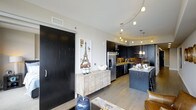 This single-floor space showcases a thoughtfully designed open-concept layout that seamlessly connects the living and kitchen areas. The kitchen is a standout feature, highlighted by sleek dark cabinetry that extends to the ceiling, creating a bold and sophisticated aesthetic. A warm metallic mosaic tile backsplash adds texture and visual interest, complementing the stainless steel appliances, including a French door refrigerator and dishwasher. The kitchen island, topped with a light-veined stone countertop, provides additional workspace and casual seating with bar-height chairs. Overhead, a combination of pendant lights and a curved track lighting system illuminates the kitchen, adding both function and style. The living area transitions naturally from the kitchen, grounded by light-toned hardwood flooring that runs throughout. A large, dramatic black barn-style sliding door serves as a striking architectural accent on one wall, offering both a design focal point and practical room division. A light wood credenza anchors the living space, while neutral walls keep the overall palette balanced and inviting.