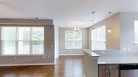 This single-floor space showcases a thoughtfully designed open-concept layout that effortlessly connects the kitchen, dining, and living areas into one cohesive living environment. The interior is finished with warm hardwood-style flooring that runs throughout, complementing the neutral beige and greige tones of the walls for a clean, welcoming aesthetic.

The kitchen features a peninsula with a light-toned granite countertop and dark cabinetry below, offering both functional workspace and a natural divider between the kitchen and the adjoining dining area. Pendant lighting above the peninsula adds a modern accent, while a stylish chandelier over the dining area provides an elegant focal point.

Multiple large windows fitted with white horizontal blinds flood the space with abundant natural light, creating an airy and open atmosphere. Views of lush greenery are visible through the windows, enhancing the sense of connection to the outdoors. White trim and baseboards throughout provide crisp contrast against the warm wall tones, adding a polished finishing touch.

The open flow of the floor plan, combined with the natural light and cohesive design elements, makes this a versatile and inviting space well-suited for both everyday living and entertaining.