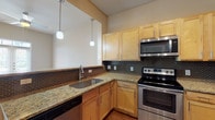 This single-floor, 1-bedroom, 1-bathroom residence offers a well-designed layout that balances comfort and functionality. The kitchen is a standout feature, equipped with stainless steel appliances including a range, over-the-range microwave, dishwasher, and refrigerator. Light maple cabinetry provides ample storage, while granite countertops and a dark mosaic tile backsplash add a refined touch. Pendant lighting illuminates the space with a warm glow, and a breakfast bar peninsula connects the kitchen to the adjacent living and dining areas. The living area features warm wood-style laminate flooring and is anchored by a set of French doors with transom windows above, flooding the room with natural light and providing access to the outdoors. The bedroom is generously sized, finished with plush neutral carpeting and painted in soft, warm tones, creating a serene retreat. The bathroom features a full soaking tub and shower combination surrounded by classic white subway tile, offering a clean and timeless aesthetic. Track lighting and ceiling fans are present throughout, and the overall neutral color palette gives the entire space a bright, inviting ambiance.