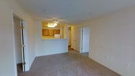 This single-floor residential unit offers a practical and comfortable layout designed for everyday living. The living area is finished with wall-to-wall carpeting in a neutral tone, creating a warm and inviting atmosphere throughout the space. A ceiling fan is centrally mounted, providing both lighting and air circulation to the room.

One of the defining features of this unit is the pass-through kitchen window, which opens the kitchen to the living area and enhances the sense of openness between the two spaces. The kitchen is equipped with warm wood-toned cabinetry and includes a refrigerator, with recessed lighting illuminating the work area from above.

Adjacent to the living space, classic six-panel white doors lead to additional rooms, offering a traditional architectural touch that complements the neutral color palette of the unit. The hallway beyond provides access to further areas of the home, suggesting a well-organized and functional floor plan.

Throughout the unit, the soft beige and cream tones of the walls and flooring contribute to a cohesive, understated aesthetic. The layout balances openness in the shared living areas with defined, private spaces accessible through the corridor, making this a well-rounded and livable single-floor unit.