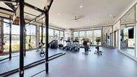 Located on Watervista Terrace in Pooler, Georgia, this generously sized single-floor fitness center offers a comprehensive workout environment designed with both functionality and aesthetics in mind. The space is defined by its expansive, open layout with durable rubber flooring throughout, providing ample room for a wide variety of exercise activities.

Large, floor-to-ceiling windows line multiple walls, flooding the interior with natural light and offering views of the surrounding greenery and neighborhood. A ceiling fan and recessed lighting complement the bright, airy atmosphere. The main workout area houses a full suite of cardio equipment, including treadmills, ellipticals, stair climbers, and stationary bikes from quality brands such as TRUE Fitness.

A dedicated strength training zone features a multi-rack dumbbell station with a broad weight range, a weight bench, plate-loaded machines, and a TRX suspension system with battle ropes for functional training. Full-length mirrors and a decorative accent wall panel enhance the strength area's visual appeal.

Glass-paneled interior walls and double doors connect to an adjacent room, adding transparency and a sense of additional space. The overall ambiance is clean, modern, and well-organized, creating an inviting environment suited for a comprehensive fitness routine.