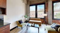 Located in the heart of downtown San Jose at S 1st Street, this one-bedroom, one-bathroom residence showcases a striking industrial-loft aesthetic throughout. Soaring concrete beam ceilings, exposed brick accent walls, and light hardwood flooring define the open living area, where oversized black-framed windows invite abundant natural light and urban views. The living room offers a generous, airy feel with dramatic ceiling height and direct sightlines to the city streetscape.

The kitchen is outfitted with rich, dark-stained wood cabinetry, black tile backsplash, dark countertops, and a full suite of stainless steel appliances, including a refrigerator, dishwasher, microwave, and range — combining style with functional efficiency.

The spacious primary bedroom features exposed brick walls, soaring ceilings with concrete beams, large windows with greenery views, soft carpeting, and an expansive wall of built-in closet storage. A dedicated office area provides additional flexibility, while two closets offer ample storage throughout.

The bathroom is thoughtfully finished with dark tile surrounds, a soaking tub and shower combination, a sleek wall-mounted vanity, and a backlit round mirror. In-unit laundry and a hallway complete this well-appointed urban residence.