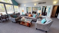 This single-story clubhouse is designed to provide a comfortable and inviting community gathering space. The architecture is defined by dramatic vaulted ceilings that rise to a prominent arched window at the front entry, flooding the interior with natural light and creating an open, airy atmosphere. Teal accent walls complement the neutral gray tones used throughout, lending the space a cohesive and contemporary aesthetic.

The main lounge area is generously sized, furnished with plush sofas and armchairs accented by teal and orange throw pillows. A glass-top coffee table serves as a central focal point, while large windows and glass doors along the perimeter offer outdoor views and additional natural light. A ceiling fan with recessed lighting overhead ensures year-round comfort. Large mirrors on one wall visually expand the space and reflect the warm, welcoming atmosphere.

Adjacent to the lounge, a dining area provides seating for groups, making the clubhouse well-suited for social gatherings. A self-serve refreshment station with a pass-through counter features a coffee maker, K-Cup carousel, and complimentary refreshments, supported by a functional kitchen with white cabinetry and a range.

The entry foyer showcases soaring ceilings, a pendant chandelier, and patterned carpet that continues throughout, tying the entire space together with a polished, well-appointed finish.