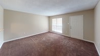 This single-floor apartment offers a practical and comfortable layout designed for efficient everyday living. The living area is generously proportioned, featuring warm brown carpeting, neutral beige walls, and a textured ceiling throughout. A large window fills the space with natural light, while a convenient pass-through window connects the living room to the kitchen, enhancing the open feel between the two areas. The kitchen is outfitted with white cabinetry, a white appliance suite including a range, and light-toned countertops, with easy-care tile flooring underfoot. A dedicated entry door opens directly into the bright living space. The bedroom is a comfortable retreat, carpeted in the same warm tones and featuring two reach-in closets for ample storage. A window brings in natural light, and a ceiling light fixture provides overhead illumination. The full bathroom includes a vanity with countertop sink, a tub/shower combination enclosed by a curtain rod, and tile flooring. A hallway connects the main living areas and includes built-in storage. Central HVAC vents are present throughout, ensuring year-round climate comfort. The overall palette of warm neutrals and consistent finishes gives the unit a cohesive, move-in-ready appeal.