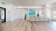 Located at 1650 Ferry Wharf Road in Kelowna, BC, this contemporary amenity lounge offers a well-appointed, open-concept recreational space with a thoughtfully designed layout spread across a single floor. Light wood-look flooring runs throughout, complementing the crisp white walls and recessed lighting that create a bright, airy atmosphere.

The space is anchored by a full-size pool table with a navy felt surface, positioned centrally atop a neutral area rug, with a dedicated cue rack nearby. Adjacent to it sits a foosball table, adding to the recreational appeal. A comfortable sectional sofa faces a wall-mounted flat-screen TV set against a warm wood-paneled feature wall, creating a relaxed lounge area accented by potted greenery.

A round dining table with upholstered chairs provides a casual seating option, while a built-in counter with overhead cabinetry adds functional utility to the space. Structural columns are seamlessly integrated into the open floor plan without disrupting the sense of flow.

Expansive windows on multiple walls flood the interior with natural light and frame impressive views of the surrounding mountains, Okanagan Lake, and the Kelowna skyline. A glass-door entry and balcony access further connect the space to its elevated surroundings, making this lounge an exceptional communal retreat.