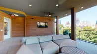 This covered outdoor terrace is designed as a sophisticated urban retreat, blending open-air living with refined architectural details. The space is framed by warm wood-paneled walls and structural columns that complement the concrete ceiling overhead, which is fitted with recessed lighting and a ceiling fan to ensure comfort in all conditions. Large open-air gaps between the columns provide an unobstructed connection to the surrounding cityscape, offering panoramic views of a downtown skyline and a lush tree canopy below, all secured by a sleek glass-panel railing along the perimeter.

The lounge area serves as the focal point of the terrace, anchored by a generously sized modular sectional sofa in neutral upholstery and paired with a slatted round coffee table. A wall-mounted flat-screen television is recessed into the wood-paneled feature wall, creating a comfortable outdoor entertainment zone. Woven accent chairs are positioned near the open balcony edge to take full advantage of the views.

A dedicated dining and kitchen zone occupies one corner of the terrace, featuring a wet bar with a sink, under-counter appliances, and pendant lighting above. Multiple dining tables surrounded by woven chairs offer ample seating for gatherings. Large-format stone tile flooring runs throughout, unifying the space with a clean, contemporary finish.