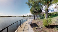 This outdoor waterfront space offers a tranquil and scenic environment centered around a paved pedestrian walkway that runs alongside a calm, expansive lake. The path is bordered by sturdy black metal railings that provide both safety and a clean, defined edge between the walkway and the water's edge. A classic slatted bench is positioned along the path, inviting visitors to pause and take in the open water views that stretch across to a tree-lined and residential far shore.

The landscape is dotted with a mix of mature palm trees and broad-canopied shade trees, lending the space a distinctly tropical character while also providing natural shelter from the sun. The grassy embankment rising gently from the path adds a soft, natural contrast to the hard surfaces of the walkway. Aquatic vegetation is visible near the water's surface in areas, adding to the natural, unspoiled feel of the shoreline.

In the distance, a bridge is visible spanning the waterway, providing a sense of the broader urban and natural context. The open sky above, typically sunny and dotted with clouds, enhances the bright and airy ambiance of the space. Overall, this waterfront walkway is a well-maintained, accessible outdoor area that blends natural scenery with functional design, ideal for leisurely strolls and quiet relaxation.