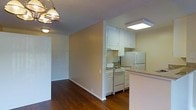 This single-floor space features an open layout that connects the living area to the kitchen, creating a cohesive and functional floor plan. The living area is finished with rich hardwood flooring that extends throughout, adding warmth to the neutral-toned walls. A classic multi-bulb chandelier provides ambient lighting to the living space, complementing the overall aesthetic.

The kitchen is efficiently designed with white upper cabinetry offering ample storage, and is equipped with white appliances including a range and a full-size refrigerator. A peninsula counter with a light-toned surface separates the kitchen from the living area, providing additional workspace and a natural transition between the two zones. A flush-mounted ceiling light fixture brightens the kitchen area directly.

The textured ceiling adds a subtle architectural detail consistent throughout the space. Crisp white baseboards and trim provide clean contrast against the warm wall tones and dark flooring. A doorway visible from the main living area suggests additional rooms or storage beyond, contributing to the overall functional layout of this well-maintained space.