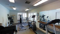 This fitness room offers a practical and comfortable environment for exercise. The space is outfitted with a selection of cardio and strength training equipment, including a treadmill, an elliptical machine, and a cable weight machine paired with a workout bench — providing a well-rounded workout experience. A large wall-mounted mirror runs along one side of the room, visually expanding the space and allowing users to monitor their form during exercise. The neutral-toned walls and carpeted flooring contribute to a clean, functional aesthetic. Multiple windows fitted with white horizontal blinds line two walls, allowing natural light to fill the room while maintaining privacy. A potted plant near the window adds a subtle touch of greenery to the otherwise utilitarian space. Framed certificates or artwork are displayed on the wall near the mirror, adding a modest decorative element. Overhead fluorescent lighting ensures the room remains well-lit at all times. The overall ambiance is straightforward and purpose-driven, designed to accommodate a variety of fitness routines in a tidy, organized setting.