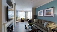 This single-floor space showcases a thoughtfully designed open-concept living area that seamlessly blends comfort and contemporary style. The living room serves as the central gathering space, highlighted by a striking teal accent wall adorned with framed artwork, which adds a bold pop of color against the neutral warm-toned walls. Crown molding runs along the ceiling, lending an elegant architectural detail throughout.

The flooring throughout is a light hardwood-style material that contributes to the bright, airy feel of the space. A fireplace with a white mantle anchors one side of the room, providing both a functional and decorative focal point. Decorative columns subtly define the transition between areas.

A generously windowed bay area at the far end of the living space floods the room with abundant natural light, featuring patterned drapes and blinds for adjustable privacy. A ceiling fan with a light fixture is installed in this sunlit nook, which accommodates additional seating. The overall ambiance is polished yet inviting, with coordinated furnishings including a neutral sofa with green accent pillows and a glass-top coffee table completing the sophisticated aesthetic.