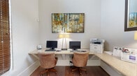 This single-floor business center is a thoughtfully designed space that balances functionality with a welcoming aesthetic. The workstation area features an L-shaped granite countertop desk that wraps around the corner, accommodating two desktop computer setups side by side, complemented by comfortable upholstered rolling chairs and a multifunction printer station. Warm-toned hardwood flooring anchors the workspace, while a large, colorful abstract painting mounted above the desk adds a vibrant artistic focal point to the otherwise clean, neutral-toned walls. Soft ambient lighting from a sleek floor lamp enhances the professional atmosphere. The adjoining lobby area makes a strong first impression with soaring ceilings, large windows dressed with wood-toned blinds that allow for adjustable natural light, and elegant tile flooring laid in a classic pattern. A statement chandelier hangs overhead, casting a warm glow over the entryway. A seating area with upholstered chairs is visible beyond, positioned beneath track lighting and tasteful wall décor, creating an inviting transitional zone. Together, these areas form a cohesive, polished environment well-suited for business operations or a community amenity space.