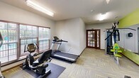 Located at 2540 Bluffs View Dr in Raleigh, NC, this dedicated fitness center offers a focused and functional space for exercise and wellness. The room is designed with an energetic aesthetic, featuring bold lime-green accent walls that create a motivating atmosphere, complemented by neutral gray walls and warm wood baseboards throughout.

The exercise room is outfitted with a well-rounded selection of equipment, including treadmills, elliptical machines, a recumbent bike, and a multi-station strength-training cable machine with lime-green accents that coordinate with the room's color scheme. A full-length mirror spans one wall, visually expanding the space and allowing users to monitor their form during workouts. A wall-mounted television is positioned for convenient viewing during cardio sessions.

Natural light filters in through multiple windows fitted with horizontal blinds, offering views of the surrounding greenery outside. Overhead fluorescent lighting ensures the room remains well-lit at all times. The floor is covered in durable patterned carpet tiles in muted yellow and gray tones, providing both comfort and practicality underfoot.

Entry is through elegant French doors with glass pane inserts, adding a refined architectural detail to the space. The room's practical layout and cohesive design make it a well-appointed fitness destination.