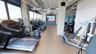 This fitness center delivers a comprehensive workout environment across a thoughtfully organized single-floor layout. The space is defined by an industrial-modern aesthetic, with exposed concrete columns, open ductwork overhead, and rubber flooring throughout. Large floor-to-ceiling windows line the perimeter, flooding the space with natural light and framing expansive city skyline views — a standout feature visible from the cardio equipment zone, where treadmills, ellipticals, stair climbers, and rowing machines are arranged to maximize the outlook.

A dedicated strength training area houses a full dumbbell rack, weight benches, cable machines, and spin bikes positioned beneath mirrored walls. A functional training zone includes a heavy bag, kettlebells, medicine balls, TRX suspension trainers, and an interactive reaction training system mounted on a vivid mural wall. A large-format video display wall serves as a focal point in the main cardio area, offering immersive visual content during workouts.

A separate studio space features Peloton bikes, mirrored walls, and additional city views, while an adjacent storage room provides organized shelving for yoga mats and accessories. The overall ambiance is energetic and modern, combining professional-grade equipment with an uplifting, light-filled environment.