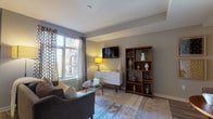 This thoughtfully designed single-floor residence offers a cohesive blend of modern aesthetics and functional living. The living room serves as the heart of the home, featuring large windows dressed with patterned drapes that invite natural light, warm wood flooring, and a comfortable seating arrangement anchored by a decorative area rug. A wall-mounted TV, a sleek white sideboard, and an open wood shelving unit provide both storage and visual interest.

The kitchen is outfitted with white upper cabinetry, dark lower cabinets, light countertops, and a full suite of stainless steel appliances, including a gas range, microwave, and dishwasher. A mobile kitchen island adds flexible prep space.

The primary bedroom is a serene retreat with access to a private balcony offering outdoor views, wood-look flooring, and warm ambient lighting. Its en-suite bathroom features a dark wood vanity, backlit mirrors, large-format tile flooring, and a glass-enclosed walk-in shower. A second well-proportioned bedroom continues the neutral, contemporary palette and is served by a full bathroom with a soaking tub and shower combination. Throughout the residence, a cohesive gray and warm-tone color scheme, clean lines, and carefully curated furnishings contribute to a polished, modern ambiance.