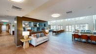 Located in Surprise, Arizona, this impressive single-floor community clubhouse offers a generously sized, thoughtfully designed gathering space that seamlessly blends elegance with functionality. The centerpiece of the facility is a soaring great room with dramatic vaulted ceilings accented by exposed wood beams and oversized globe pendant lights. Large floor-to-ceiling windows and glass doors flood the space with abundant natural light while providing views of the surrounding outdoor areas. Light-toned hardwood flooring in a herringbone pattern flows throughout, lending warmth and continuity to the open layout. A full kitchen island with bar seating anchors the dining area, which features multiple communal tables with rich amber upholstered chairs, ideal for shared meals and events. Adjacent to the main hall, a welcoming lobby with a distinctive botanical chandelier and herringbone wood flooring provides an elegant entry point. Comfortable lounge areas are thoughtfully arranged with plush sofas, accent chairs in cool blues and warm neutrals, and built-in shelving with blue inset cubbies. Glass-walled private meeting rooms offer flexible spaces for gatherings or work. A dedicated game room, complete with a billiard table and carpeted seating areas, rounds out the amenities. The overall ambiance is modern, airy, and inviting.