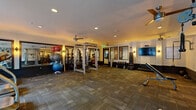 This fitness center offers a generously sized, single-floor workout space designed to support a wide range of exercise routines. The room is finished with durable, patterned carpet tiles throughout and illuminated by recessed ceiling lights complemented by warm wall sconces, creating a welcoming yet functional atmosphere. Ceiling fans provide additional air circulation across the space.

The cardio zone is positioned along one wall of large floor-to-ceiling windows, offering views of lush outdoor greenery and a balcony area, filling the space with natural light. This section houses multiple treadmills, elliptical machines, and a stationary bike, all arranged to take advantage of the scenic outlook.

The strength training area is centrally located and anchored by a full power rack with a barbell and weight plates, ideal for compound lifts. A dedicated free weights section features a rack of dumbbells and a colorful assortment of medicine balls and stability balls of various sizes, supporting functional and core training. An adjustable bench and cable machine add further versatility.

Mirrored wall panels line one side of the room, visually expanding the space and allowing users to monitor their form during workouts. A wall-mounted flat-screen television is also present. The overall design balances practicality with an inviting ambiance, making this fitness center a comprehensive and comfortable training environment.