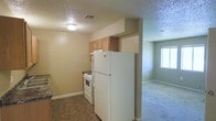 This single-floor residential unit offers a practical and straightforward layout designed for comfortable everyday living. The kitchen is equipped with full-size white appliances, including a refrigerator and an electric range, complemented by light wood upper cabinetry that provides ample storage. Dark countertops offer a subtle contrast to the neutral, warm-toned walls, while durable vinyl flooring grounds the space. A textured ceiling adds a classic finishing touch throughout. The kitchen flows openly into the adjacent living area, which features soft wall-to-wall carpeting underfoot, creating a welcoming transition between functional and relaxing spaces. A window in the living area allows natural light to filter in, brightening the room and contributing to an airy feel. The overall design is clean and utilitarian, with a neutral color palette throughout that makes the space easy to personalize. Flush-mounted ceiling lighting ensures consistent illumination in the kitchen, while the open connection between rooms enhances the sense of space and continuity across this single-level layout.