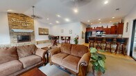 This single-floor community space at Independence Oaks offers a thoughtfully designed layout that balances comfort and functionality. The expansive living room serves as the heart of the space, anchored by a dramatic floor-to-ceiling stacked stone fireplace with a classic white mantel and raised stone hearth. Vaulted ceilings with ceiling fans add to the open, airy atmosphere, while recessed lighting and large windows flood the room with natural light. Plush seating arrangements and neutral carpet create an inviting lounge environment, complemented by framed wall art and decorative greenery. Glass-panel entry doors connect the living area to adjacent spaces, offering visibility and a sense of openness. A secondary seating area near the windows provides a quiet retreat with views to the outdoors. The kitchen is well-appointed with rich cherry wood cabinetry reaching to the ceiling, dark granite-style countertops, and black appliances including a refrigerator and range. A standout feature is the stone-clad peninsula breakfast bar with bar stool seating, echoing the stonework found in the living room and tying the two spaces together cohesively. Tile flooring in the kitchen provides a durable, clean finish that transitions smoothly throughout the functional areas of this welcoming community gathering space.