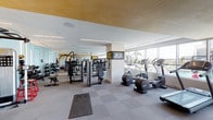 This fitness center offers a comprehensive workout environment designed with both functionality and aesthetics in mind. The space is characterized by a warm wood-paneled ceiling accented by recessed linear lighting, which contrasts with crisp white walls to create a clean, modern atmosphere. Large floor-to-ceiling windows line one side of the facility, flooding the interior with natural light and providing views of an outdoor pool area and the surrounding city skyline—an motivating backdrop for any workout.

The cardio zone features multiple treadmills and elliptical machines positioned directly in front of the expansive windows, allowing users to enjoy the outdoor scenery while exercising. A wall-mounted flat-screen TV is centrally positioned for easy viewing throughout the space.

The strength training area is well-appointed with a variety of cable machines, weight benches, and selectorized equipment arranged to maximize usability. A full-length mirror wall extends across one section, visually expanding the space while providing functional feedback during workouts. A dedicated free weights section includes a multi-tiered dumbbell rack, medicine balls, and plyo boxes. The flooring throughout consists of a durable, checkered rubber tile in neutral tones, providing cushioning and traction across all exercise zones.