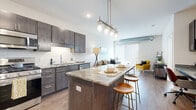 This well-designed single-floor residence offers a thoughtful layout that balances functionality and modern aesthetics. The open-concept kitchen is a standout feature, equipped with dark espresso cabinetry, granite countertops, a full suite of stainless steel appliances including a gas range, microwave, dishwasher, and refrigerator, and a white subway tile backsplash. A central kitchen island with stone countertops and pendant lighting above serves as both a prep space and a casual dining area with bar seating. Wood-style flooring flows seamlessly from the kitchen into the living area, which is bright and airy thanks to a large sliding glass door fitted with vertical blinds. An exposed spiral duct adds an industrial design accent to the living space. The bedroom features plush carpeting, generous natural light from a wide window with blinds, and neutral wall tones that create a calm, restful atmosphere. The bathroom is finished with a dark wood-tone vanity, white quartz countertop, a large mirror, and coordinating hardware, complemented by wood-style flooring. Throughout the residence, the design palette of dark wood tones, neutral walls, and brushed nickel accents creates a cohesive and contemporary living environment.