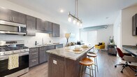 This well-designed single-floor residence offers a thoughtful layout that balances functionality and modern aesthetics. The open-concept kitchen is a standout feature, equipped with dark espresso cabinetry, granite countertops, a full suite of stainless steel appliances including a gas range, microwave, dishwasher, and refrigerator, and a white subway tile backsplash. A central kitchen island with stone countertops and pendant lighting above serves as both a prep space and a casual dining area with bar seating. Wood-style flooring flows seamlessly from the kitchen into the living area, which is bright and airy thanks to a large sliding glass door fitted with vertical blinds. An exposed spiral duct adds an industrial design accent to the living space. The bedroom features plush carpeting, generous natural light from a wide window with blinds, and neutral wall tones that create a calm, restful atmosphere. The bathroom is finished with a dark wood-tone vanity, white quartz countertop, a large mirror, and coordinating hardware, complemented by wood-style flooring. Throughout the residence, the design palette of dark wood tones, neutral walls, and brushed nickel accents creates a cohesive and contemporary living environment.