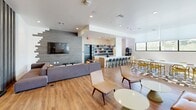 This single-floor amenity lounge offers a generously sized, open-plan layout thoughtfully divided into distinct functional zones through the use of sleek vertical slatted partitions. The warm hardwood flooring flows continuously throughout, tying the various areas together while reinforcing the space's contemporary aesthetic.

The recreation zone is well-equipped, featuring a ping-pong table, a pool table, and a full-length shuffleboard table, all set against clean white walls accented with framed artwork. Built-in cabinetry provides dedicated storage for cues and game accessories.

Adjacent to the game area, a comfortable lounge zone offers a large sectional sofa with colorful accent pillows arranged around low round coffee tables, paired with a wall-mounted flat-screen TV and a striking textured accent wall. Woven accent chairs add warmth and variety to the seating arrangement.

The dining and café area is bathed in natural light from a continuous row of large windows, featuring a mix of high-top bar tables with modern stools and a casual dining table with vibrant yellow chairs set atop a patterned tile-inset floor. A well-appointed bar counter with open shelving, pendant lighting, and a coffee station completes this zone, making it ideal for casual gatherings.