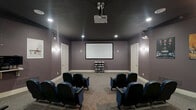 Located at 6320 Clock Tower Dr in Walbridge, Ohio, this purpose-built private screening room delivers an authentic cinema experience within a compact, well-appointed space. The room is finished with deep mauve-gray walls and a dark ceiling that absorb light and enhance contrast, creating an immersive viewing atmosphere. Recessed ceiling lights and wall sconces provide soft, controlled ambient illumination when needed.

The centerpiece of the room is a large wall-mounted projection screen paired with a ceiling-mounted digital projector, offering a true theater-quality viewing setup. Multiple rows of blue and black upholstered theater-style seats with fold-down armrests are arranged in a slightly curved layout, ensuring clear sightlines from every position. The seating comfortably accommodates a private group, making it ideal for screenings and events.

Along the walls, framed classic movie posters — including titles such as Goodfellas, Braveheart, The Lord of the Rings, and Forrest Gump — add character and reinforce the cinematic theme. A media storage rack near the screen holds an organized collection of physical media. Soft carpet throughout contributes to acoustic comfort and a refined aesthetic. Two white panel doors provide access points, and the overall design prioritizes both function and atmosphere.