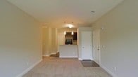 This single-floor residence features an open-concept design that seamlessly connects the living area to the kitchen. The living space is finished with wall-to-wall neutral carpeting, contributing to a warm and comfortable atmosphere, while the entryway transitions to hard flooring near the front doors. Two white panel doors are positioned near the entry, likely serving as closet and main entry access points.

The kitchen is defined by a raised breakfast bar peninsula with granite-style countertops, providing both a functional workspace and an informal dining option. Dark cabinetry lines the upper walls of the kitchen, offering ample storage, and a built-in microwave or range hood is visible above the cooking area. Pendant lights hang above the peninsula, adding focused task lighting and a decorative touch.

The ceilings are smooth and finished in a neutral tone, with recessed vents integrated cleanly into the layout. A doorway along the left wall leads further into the unit, suggesting additional rooms beyond the main living and kitchen area. The overall palette of beige walls, neutral carpeting, and dark kitchen accents creates a balanced, move-in-ready aesthetic.
