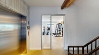 This modern fitness facility is thoughtfully designed across a single floor, offering a generous and well-organized layout divided into distinct functional zones. The main cardio and strength training area features polished concrete flooring, exposed wooden ceiling beams, and large windows that invite abundant natural light while providing views of the surrounding exterior. Ceiling fans are strategically placed throughout to maintain airflow and comfort during workouts. The strength zone is equipped with a multi-functional cable machine, free weights, and a dedicated dumbbell rack set against warm wood-paneled accent walls, which add a natural, inviting texture to the space. A separate studio room offers a versatile open floor plan ideal for group fitness or yoga, distinguished by a tray ceiling painted in a soft sage green, bold botanical-print wallcovering, full-length mirrors, and glass-panel doors that open to the outdoors. The entry area features warm wood ceiling planks, a staircase with rich hardwood treads and black iron railings, and an elevator with white tile surround for convenient access. Throughout, the design blends contemporary clean lines with natural wood elements, creating a welcoming and functional environment that encourages active living.