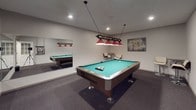 Located at 301 T St in Tumwater, Washington, this dedicated game room is designed for recreation and social enjoyment. The centerpiece of the space is a full-size pool table with a vibrant teal felt surface, complemented by a striking overhead billiard light fixture featuring bold red shades and a decorative ball rack — providing both functional illumination and a classic game room aesthetic. Recessed lighting throughout the smooth, neutral-toned ceiling ensures the space is well-lit from every angle. A wall-mounted cue rack keeps billiard cues neatly organized and easily accessible, while a large wall mirror expands the sense of space and adds a dynamic visual element to the room. A bistro-style seating area with two adjustable bar stools and a round high-top table offers a comfortable spot for spectators or casual conversation. The flooring is covered in durable, low-pile carpet in a neutral gray tone, providing comfort underfoot and reducing noise. Decorative artwork on the walls adds personality and visual interest to the otherwise clean, minimalist design. The room is accessed through a paneled door with glass insets, contributing to a polished and cohesive interior finish throughout this inviting recreational space.