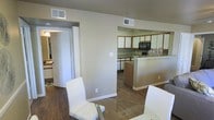 This single-floor space showcases an open-concept design that connects the dining area and kitchen in a cohesive, functional layout. The dining area features a glass-top round table with modern white chairs, providing a clean and contemporary feel. Warm hardwood-style flooring runs throughout, adding a sense of continuity and warmth to the space.

The kitchen is positioned adjacent to the dining area and is defined by a peninsula with medium-toned wood cabinetry at the base, complemented by white upper cabinets. Black appliances, including a microwave and what appears to be a range, are integrated into the cabinetry, providing a functional contrast to the lighter elements. The countertops offer ample preparation space, and the layout promotes an efficient workflow.

Neutral beige-toned walls keep the space feeling bright and versatile. A doorway visible on the left side leads to an additional area, suggesting further rooms or utility spaces beyond the main living zone. Ceiling-mounted HVAC vents are distributed throughout, ensuring consistent climate control. The overall ambiance is clean, practical, and well-suited for everyday living.
