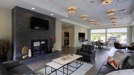 This well-appointed single-floor space combines modern design with functional comfort across several interconnected zones. The focal point of the main lounge area is a striking floor-to-ceiling dark stone fireplace, which anchors the room and adds a dramatic, contemporary touch. A large flat-screen television is mounted above the fireplace, making the space ideal for both relaxation and entertainment. The lounge is furnished with sleek dark leather seating and a clean-lined coffee table set, all resting atop a neutral area rug over rich dark hardwood-style flooring. Overhead, a drop ceiling with recessed lighting and warm flush-mount fixtures provides a layered, inviting glow throughout the space. Beyond the lounge, a kitchenette area is visible in the background, featuring dark cabinetry, a bar-height counter with seating, and essential appliances — making it a convenient hub for casual gatherings. Adjacent to the kitchenette, a dining area benefits from a large window that floods the space with natural light and offers views of lush greenery outside. The open-concept layout promotes a seamless flow between the lounge, kitchen, and dining zones, creating a versatile and welcoming environment suited for social interaction and community use.
