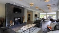 This well-appointed single-floor space combines modern design with functional comfort across several interconnected zones. The focal point of the main lounge area is a striking floor-to-ceiling dark stone fireplace, which anchors the room and adds a dramatic, contemporary touch. A large flat-screen television is mounted above the fireplace, making the space ideal for both relaxation and entertainment. The lounge is furnished with sleek dark leather seating and a clean-lined coffee table set, all resting atop a neutral area rug over rich dark hardwood-style flooring. Overhead, a drop ceiling with recessed lighting and warm flush-mount fixtures provides a layered, inviting glow throughout the space. Beyond the lounge, a kitchenette area is visible in the background, featuring dark cabinetry, a bar-height counter with seating, and essential appliances — making it a convenient hub for casual gatherings. Adjacent to the kitchenette, a dining area benefits from a large window that floods the space with natural light and offers views of lush greenery outside. The open-concept layout promotes a seamless flow between the lounge, kitchen, and dining zones, creating a versatile and welcoming environment suited for social interaction and community use.