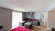 This single-floor residence offers a well-appointed living area designed with a contemporary aesthetic. The living room serves as the central gathering space, featuring a large sectional sofa in a warm gray upholstery, accented by bold red throw pillows that add a vibrant contrast to the neutral color scheme. A matching ottoman provides additional seating and functionality. A vivid pink area rug anchors the seating arrangement, introducing a striking pop of color against the light wood-style flooring that runs throughout the space.

The room is brightened by a full-width sliding glass door dressed with vertical blinds, allowing for generous natural light and providing access to an outdoor area. The textured popcorn ceiling and neutral gray walls create a cohesive backdrop, while a paneled accent wall adds subtle architectural interest. A pair of large black-and-white elephant canvas prints serves as a bold focal point above the sofa, lending an artistic and dynamic character to the space. A doorway visible from the living area leads to an additional room, suggesting a functional and connected floor plan throughout this single-level home.
