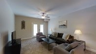 This single-floor space offers a welcoming and functional layout designed for comfortable everyday living. The living room serves as the heart of the space, finished with plush wall-to-wall carpeting that adds warmth underfoot. A ceiling fan with an integrated light fixture is centrally mounted, providing both airflow and ambient lighting throughout the room. The space is bathed in natural light, courtesy of a set of double French doors that open to the outside, creating a seamless connection between the interior and the outdoors. Neutral-toned walls provide a clean, versatile backdrop that complements the understated décor. The living area is thoughtfully arranged with a dark, cushioned sofa sectional, a coordinating armchair, and a sleek rectangular coffee table, offering ample seating for relaxing or entertaining. Decorative accents, including wall art and accent pillows, lend a lived-in, curated feel to the room. The overall ambiance is calm and inviting, with a design sensibility that balances practicality with comfort, making it a versatile space suited to a variety of lifestyles.