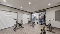 Located at 11245 West Rd in Houston, TX, this fitness center is a comprehensively outfitted exercise facility designed to support a wide range of workout styles. The space is divided into two distinct zones that together create a dynamic and functional training environment. The cycling studio features multiple stationary spin bikes arranged on smooth wood-look flooring, complemented by a wall-mounted flat-screen TV and recessed lighting overhead — ideal for group cycling classes or individual cardio sessions. The adjoining functional training area showcases a striking herringbone-patterned tile floor and is anchored by an overhead rig system supporting a heavy punching bag and TRX suspension trainers. This zone is further equipped with wall-to-wall storage racks holding an assortment of medicine balls, kettlebells, and battle ropes, alongside additional strength and conditioning equipment. Crown molding, neutral gray walls, and recessed lighting throughout lend a clean, modern aesthetic to the entire facility. Glass-paned doors connect the training area to the exterior, allowing natural light to filter in. The overall design balances functionality with a polished, inviting ambiance suitable for fitness enthusiasts of all levels.