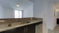 This single-floor space showcases a thoughtfully designed kitchen that serves as a functional centerpiece. The kitchen features granite countertops with a natural speckled pattern, complemented by rich dark wood cabinetry equipped with sleek bar-pull hardware. A stainless steel double-basin sink is seamlessly integrated into the countertop, accompanied by a matching stainless steel dishwasher, reflecting a cohesive modern aesthetic. A granite backsplash ties together the countertop and cabinetry, adding visual continuity throughout the kitchen space.

The kitchen opens toward an adjacent living area, which benefits from a large window that invites generous natural light into the space. The window, framed by a clean white surround, offers a view to what appears to be an outdoor balcony or exterior railing, suggesting access to outdoor space. The crisp white walls and ceilings throughout contribute to a bright, airy atmosphere. Flush-mount ceiling lighting provides additional illumination. The overall design balances practicality with clean, contemporary finishes, making for a functional and visually cohesive living environment.