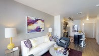 This well-appointed single-floor residence offers a seamless blend of modern design and functional living. The open-concept layout connects the living area directly to the kitchen, creating an inviting and cohesive flow throughout the space. The living room is anchored by a crisp white sofa accented with decorative throw pillows in purple and cream tones, complemented by a charcoal accent chair and a sleek round coffee table. A bold, colorful abstract artwork serves as a striking focal point on the smooth grey feature wall, adding a vibrant artistic touch to the room. Warm-toned table lighting enhances the welcoming ambiance. The flooring transitions from rich hardwood to plush area rugs, adding texture and warmth underfoot. The kitchen is visible from the living area and features dark cabinetry, stainless steel appliances including a refrigerator and range, and track lighting overhead that illuminates the workspace efficiently. The neutral grey wall palette throughout provides a versatile backdrop that allows the furnishings and artwork to stand out. Clean lines, contemporary finishes, and a thoughtful use of color give this space a polished, modern character ideal for everyday living and entertaining.