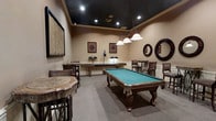 This dedicated game room is thoughtfully designed for entertainment and social gatherings. The space is anchored by a full-size pool table with elegant carved wooden legs and a classic teal felt surface, positioned beneath a trio of pendant lights that provide focused illumination. A ping pong table sits toward the rear of the room, complemented by additional task lighting for gameplay. The warm, neutral-toned walls are adorned with decorative framed artwork and circular mirrors, adding visual interest and a refined character to the space. High bar-height tables with wicker-style stools are arranged along the perimeter, offering comfortable seating for spectators and socializing. The dark-painted ceiling with recessed lighting creates a dramatic contrast against the lighter walls, lending the room a sophisticated, lounge-like ambiance. Plush carpeting underfoot adds warmth and comfort throughout. Decorative wall hangings, including a textured panel piece, further enhance the room's polished aesthetic. Overall, this game room strikes a balance between functional recreation space and stylish interior design, making it well-suited for both casual play and entertaining guests.