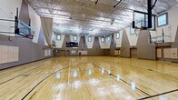 Located at 5009 Park Central Drive in Orlando, Florida, this expansive indoor sports facility offers a generously sized, single-floor gymnasium designed to accommodate serious athletic activity. The space is dominated by a gleaming, polished hardwood floor marked with multiple full and cross-court basketball configurations, allowing for simultaneous or segmented use of the floor.

Soaring ceilings supported by an open steel truss structure give the gymnasium an impressive sense of vertical volume, while rows of clerestory windows positioned high along all four walls invite abundant natural light, reducing reliance on the overhead fluorescent and pendant lighting system. The walls feature a striking geometric design, with bold triangular gray panels punctuated by the window openings, lending a modern and dynamic aesthetic to the interior.

Multiple regulation basketball hoops — both fixed and wall-mounted, adjustable units — line the perimeter, supporting multi-court play. A full electronic scoreboard is mounted prominently on one end wall for competitive game use. Retractable divider curtains allow the facility to be partitioned into separate playing zones as needed. Padded wall protectors are installed at lower wall sections to enhance player safety. Multiple access doors provide convenient entry and exit points throughout the space.