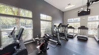 The fitness center at Bridle Creek is a thoughtfully designed workout facility that provides residents with a comprehensive exercise environment. The space features soaring ceilings that create an open, airy atmosphere, complemented by large windows along multiple walls that flood the room with natural light and offer views of the surrounding greenery. The walls are finished in a neutral gray palette, contributing to a clean and modern aesthetic.

The cardio zone is well-stocked with multiple treadmills, elliptical trainers, a stationary bike, and a rowing machine, all arranged to maximize space and usability. A dedicated strength training area features a cable weight machine, a dumbbell rack with a full-length mirror, weight benches, and stability balls, offering a complete resistance workout experience. Rubber flooring throughout the facility provides comfort and safety during exercise.

Ceiling fans help maintain airflow throughout the space, while multiple wall-mounted flat-screen televisions allow users to stay entertained during their workouts. A water fountain is conveniently located near the exit, and paper towel dispensers are available for equipment maintenance. A glass-paned door provides direct access to the outdoor area. Overall, the fitness center at Bridle Creek is a bright, well-appointed space designed to support a wide range of fitness goals.