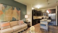 This single-floor space showcases a thoughtfully designed open-concept layout that seamlessly connects the living and kitchen areas, creating a cohesive and inviting atmosphere. The living area features warm hardwood-style flooring and is anchored by a bold sage green accent wall adorned with large-scale artwork, adding a striking visual focal point to the room. A plush neutral-toned sofa, accent side table, and layered décor elements contribute to the space's comfortable yet polished character.

The kitchen is a standout feature, outfitted with rich dark espresso cabinetry that extends to the ceiling, providing ample storage. A textured mosaic tile backsplash adds depth and dimension, while the granite countertops and stainless steel appliances — including a built-in microwave and refrigerator — speak to the kitchen's functional elegance. A breakfast bar with sleek bar seating bridges the kitchen and living area, making the space ideal for both everyday living and entertaining.

Warm, ambient lighting throughout enhances the inviting atmosphere, with a modern overhead fixture above the kitchen adding a refined touch. The open flow of the layout, combined with quality finishes and a cohesive color palette of warm neutrals and deep tones, gives this space a contemporary yet comfortable feel.