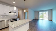 Located at 3601 Cross Hill Dr in Madison, WI, this single-floor unit offers a thoughtfully designed open-concept layout that connects the kitchen and living areas seamlessly. The kitchen is well-appointed with white shaker-style cabinetry extending to the ceiling, a subway tile backsplash, granite countertops, a central island with pendant lighting overhead, and a full suite of stainless steel appliances including a refrigerator, range, dishwasher, and microwave. Wood-look hard flooring runs throughout the main living areas, creating a cohesive and easy-to-maintain surface. The spacious living area is brightened by a sliding glass door that opens to an outdoor view of greenery, allowing ample natural light to fill the space. The bedroom features soft carpeting, a generously sized reach-in closet with dual hanging rods, and a window overlooking the surrounding landscape. Additional carpeted rooms provide flexible living options. The bathroom is finished with a granite-topped vanity, a tub-shower combination with grab bars, wood-look flooring, and clean white walls, contributing to the unit's consistently modern and neutral aesthetic throughout.