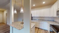 This single-story home showcases a thoughtfully designed open-concept floor plan that seamlessly connects the kitchen and living areas. The kitchen is a standout feature, outfitted with crisp white shaker-style cabinetry extending to the ceiling, speckled granite countertops, and a full suite of stainless steel appliances including a side-by-side refrigerator, electric range, and built-in microwave. A peninsula with an undermount sink and decorative seeded-glass pendant lights serves as a natural divider between the kitchen and the expansive living space. Light wood-look flooring flows throughout the main living areas, complementing the neutral warm-beige wall tones. The open living room offers generous proportions, enhanced by a ceiling fan and access to additional hallways and rooms. Bedrooms feature plush carpeting and ceiling fans, with at least one room benefiting from a large window that invites natural light. The bathroom maintains the home's cohesive aesthetic with a granite-topped vanity, white cabinetry, a wall mirror with contemporary lighting, and a tub-shower combination. Throughout, recessed lighting and clean white trim provide a polished, move-in-ready finish.