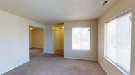 This single-floor space presents a straightforward, functional layout with an open feel throughout. The main living area is finished with wall-to-wall carpeting in a neutral beige tone, complemented by light-colored walls that contribute to a bright and airy atmosphere. A textured popcorn ceiling runs throughout, adding a classic residential character to the space.

A window fitted with horizontal blinds allows natural light to filter into the main room, providing a connection to the outdoors while maintaining privacy. The open archway visible from the main room leads into an adjacent space, also carpeted, suggesting a seamless flow between living areas. Ceiling-mounted light fixtures provide additional illumination in the connecting room.

A hallway visible from the main living area leads to what appears to be a closet with bi-fold doors, offering built-in storage solutions. The clean white trim and baseboards throughout provide a crisp contrast against the neutral wall tones. The overall layout is practical and cohesive, with each area connecting naturally to the next, making the space feel organized and easy to navigate.
